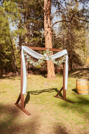 A rustic wooden wedding arch decorated with white fabric and floral accents stands in a pine forest setting with a wine barrel.