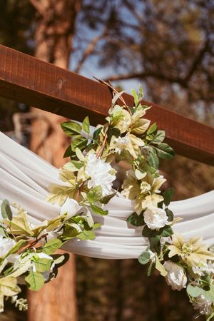 White and cream floral arrangement adorning draped fabric on wooden wedding arch detail.