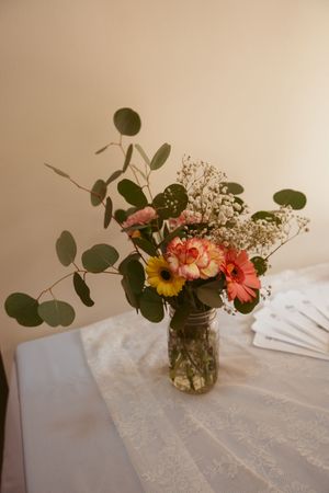 Fresh floral arrangement with gerbera daisies and eucalyptus leaves in a mason jar on a white tablecloth.