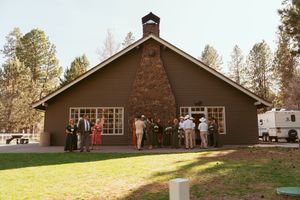 Brown rustic chapel with guests gathered outside for wedding ceremony in wooded setting.