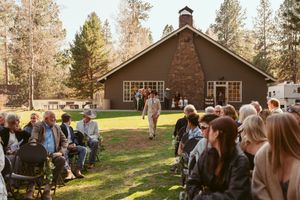 An outdoor wedding ceremony takes place in front of a brown lodge-style building surrounded by tall pine trees.