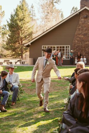 A guest in a tan suit joyfully runs during an outdoor celebration at a rustic lodge venue surrounded by pine trees.
