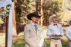 Two men in beige suits and cowboy hats stand at a rustic outdoor wedding altar.