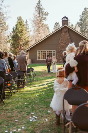 Outdoor wedding ceremony taking place on a lawn in front of a quaint chapel surrounded by tall pine trees.