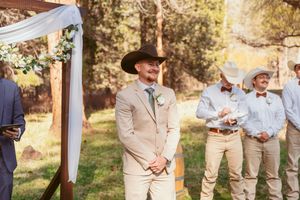Wedding ceremony setup with a simple white fabric arch and groomsmen in beige suits outdoors.