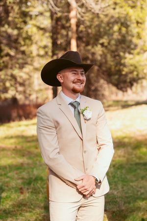 Groom in beige suit and black cowboy hat smiles while standing outdoors in rustic venue setting.
