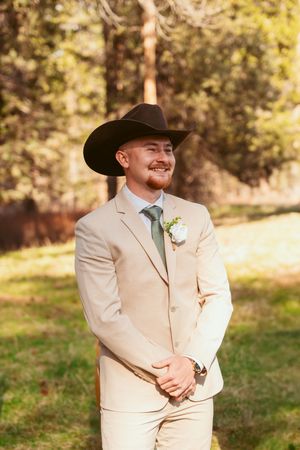 Western-style groom in beige suit and black cowboy hat smiles during outdoor wedding.
