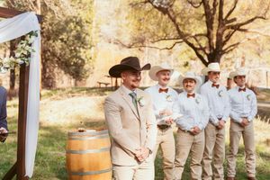 Wedding ceremony at rustic outdoor venue with groomsmen lined up wearing beige and white attire.