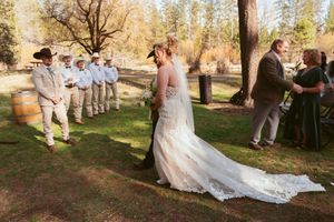 A wedding ceremony unfolds on a sunlit lawn with groomsmen in blue and guests gathered under tall trees.