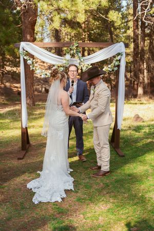 A wedding ceremony unfolds beneath a draped white arch in a serene forest setting.