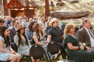 Wedding guests seated in folding chairs at outdoor forest ceremony venue.