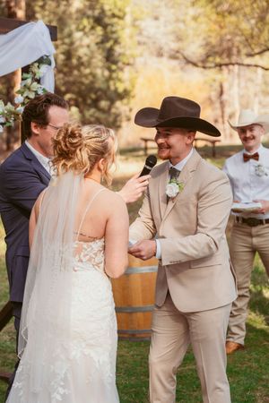 A wedding ceremony at an outdoor rustic venue with a wine barrel altar and the groom wearing a cowboy hat and tan suit.