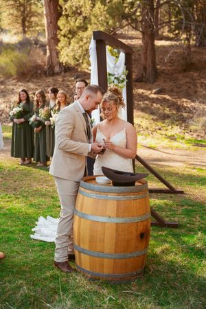 Couple performs unity ceremony at rustic outdoor wedding with wine barrel and bridesmaids in green dresses.