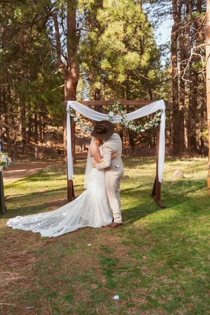 Couple embraces under white wedding arch in sunlit pine forest setting.