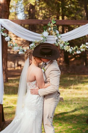 Couple embraces under a floral-decorated wedding arch with white draping in a rustic outdoor setting.