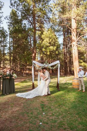 Outdoor wedding ceremony in a pine forest with decorated arch and guests on a grassy clearing.