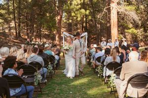 Outdoor forest wedding ceremony with guests seated on white chairs under towering pine trees.