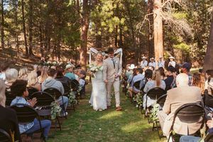 Forest wedding ceremony with guests seated on folding chairs among tall pine trees.