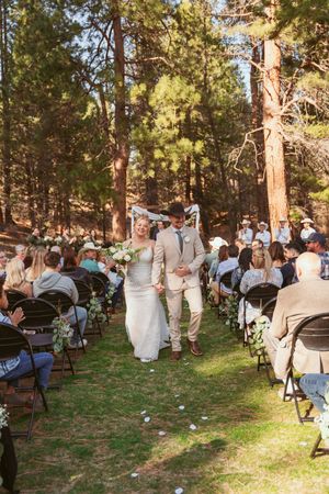 A wedding ceremony in a forest setting with guests seated on folding chairs as a couple walks down the aisle.