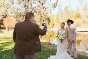 A wedding ceremony takes place on a sunny autumn day by a pond with fall foliage in the background.