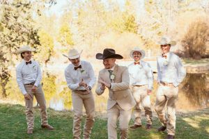 Groomsmen in cowboy hats and light colored attire posing playfully outdoors during a western-themed wedding.