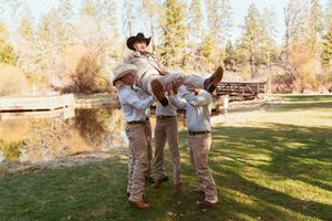 Groomsmen wearing khaki pants and white shirts playfully lift someone in a scenic outdoor setting with autumn trees.