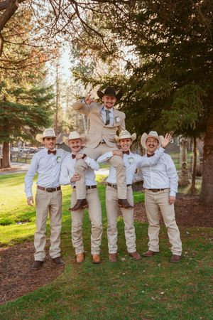 Group in western attire and cowboy hats posing playfully on grass with trees in background.