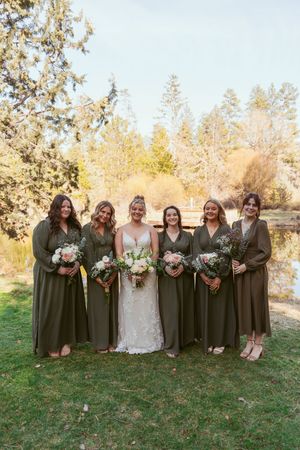 Bridesmaids in olive green dresses pose outdoors with floral bouquets against an autumn backdrop.