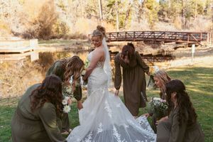 A wedding party helps adjust the delicate lace train of a bridal gown near a rustic bridge.