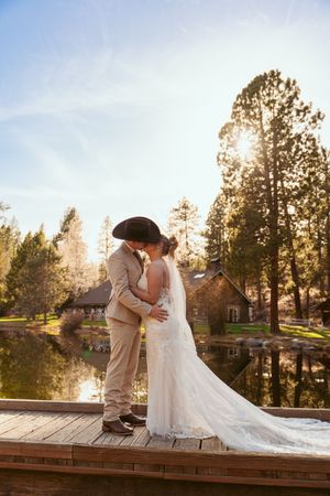A romantic sunset wedding portrait of a couple embracing on a wooden dock with tall pine trees and a lake in the background.