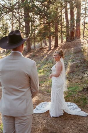 A romantic forest wedding scene with a couple in tan suit and white lace dress among tall pine trees at sunset.
