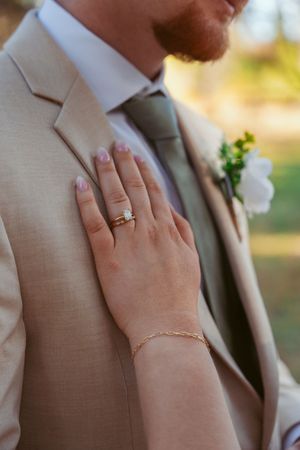 Close up of a hand wearing an engagement ring resting on a tan suit jacket with a white floral boutonniere.