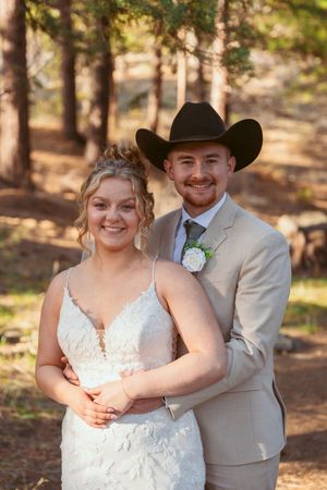 A rustic western-themed wedding couple poses in a wooded setting wearing a lace gown and beige suit with cowboy hat.