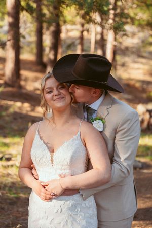 A romantic wedding couple embraces in a wooded setting with sunlit trees in the background.