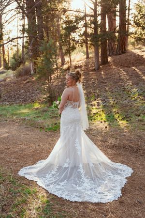 A wedding dress with intricate lace detailing and long train photographed in a sunlit forest setting.