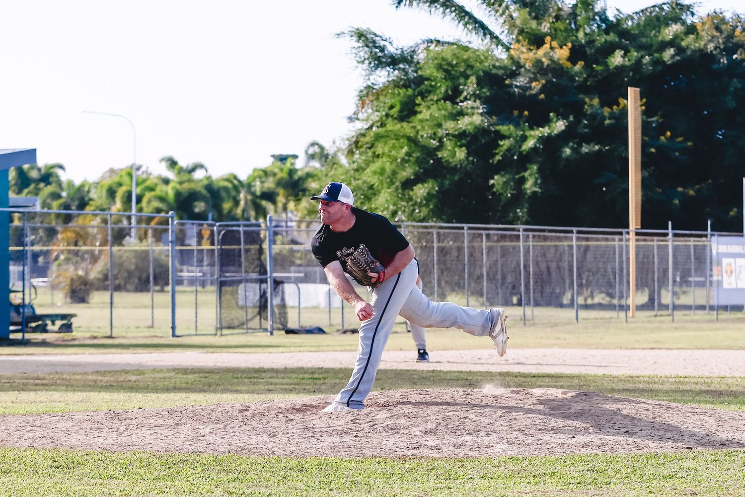 Baseball pitcher in dark uniform follows through on delivery during outdoor game on dirt mound at park field.