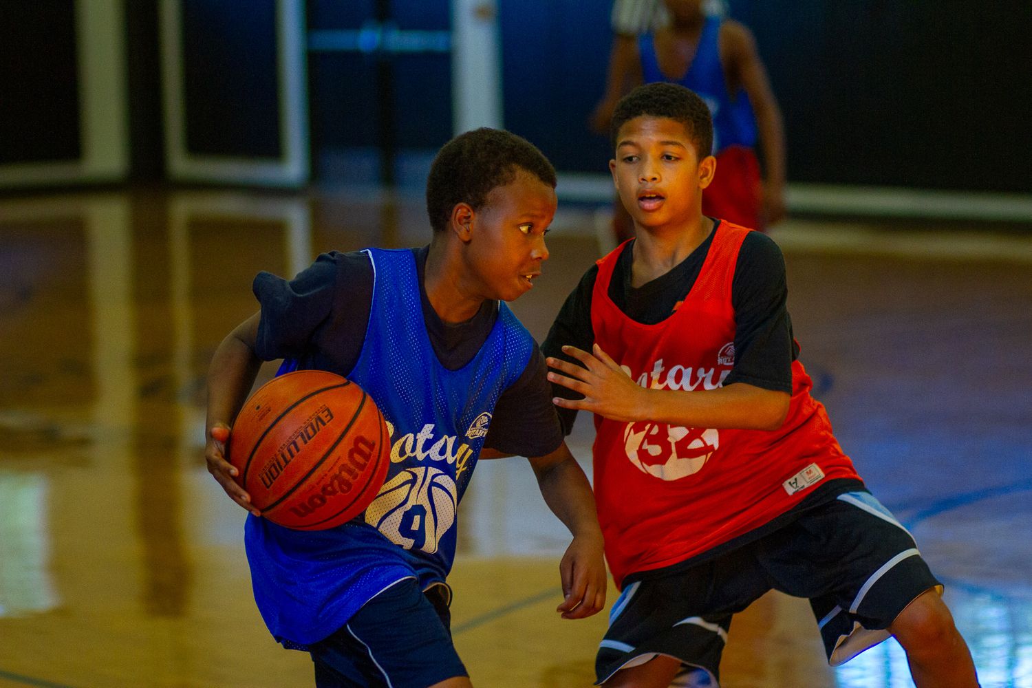 Two young basketball players compete during an indoor youth basketball game, one dribbling in blue and one defending in red.