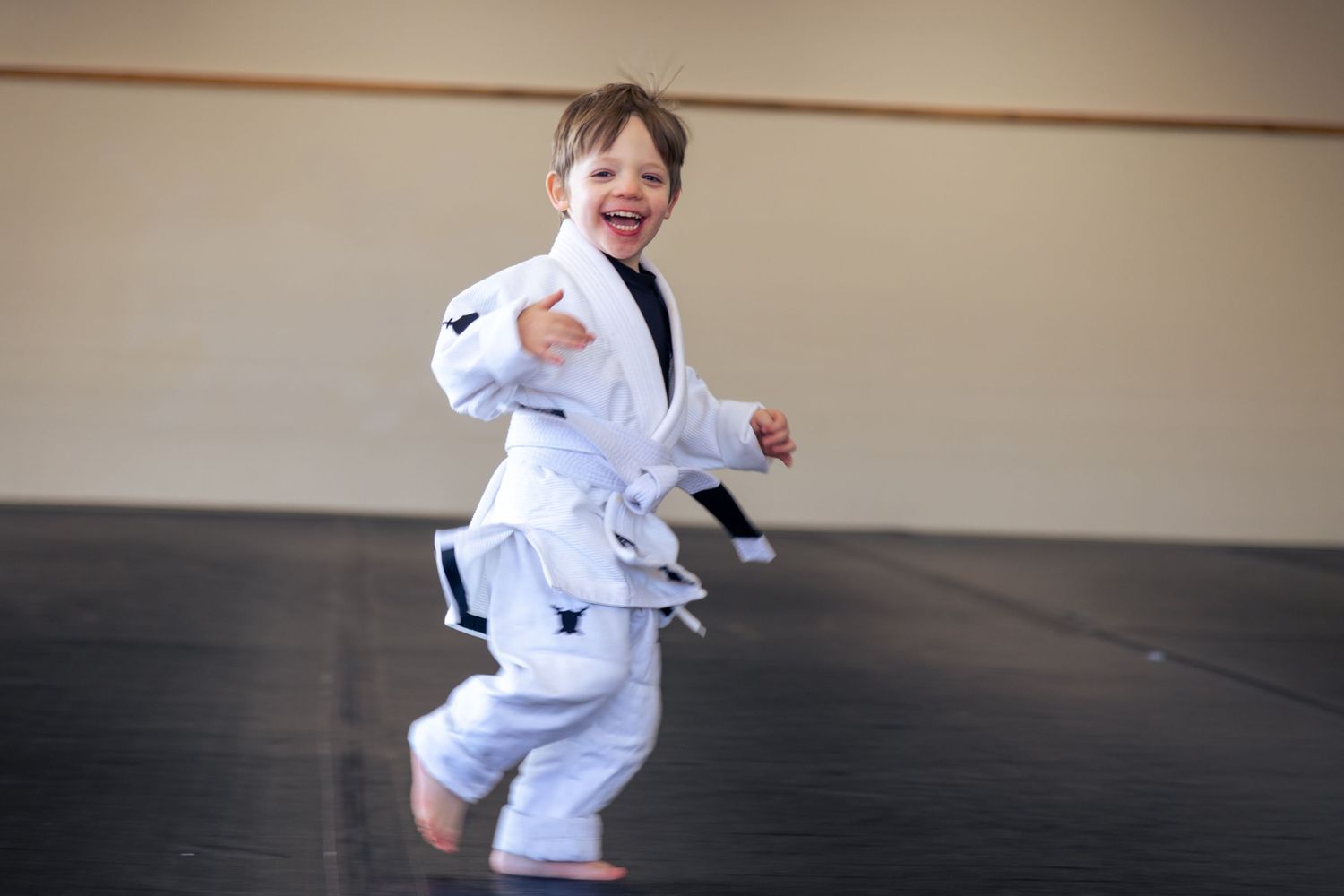 Happy toddler in white karate gi practicing martial arts on a black mat in a dojo.