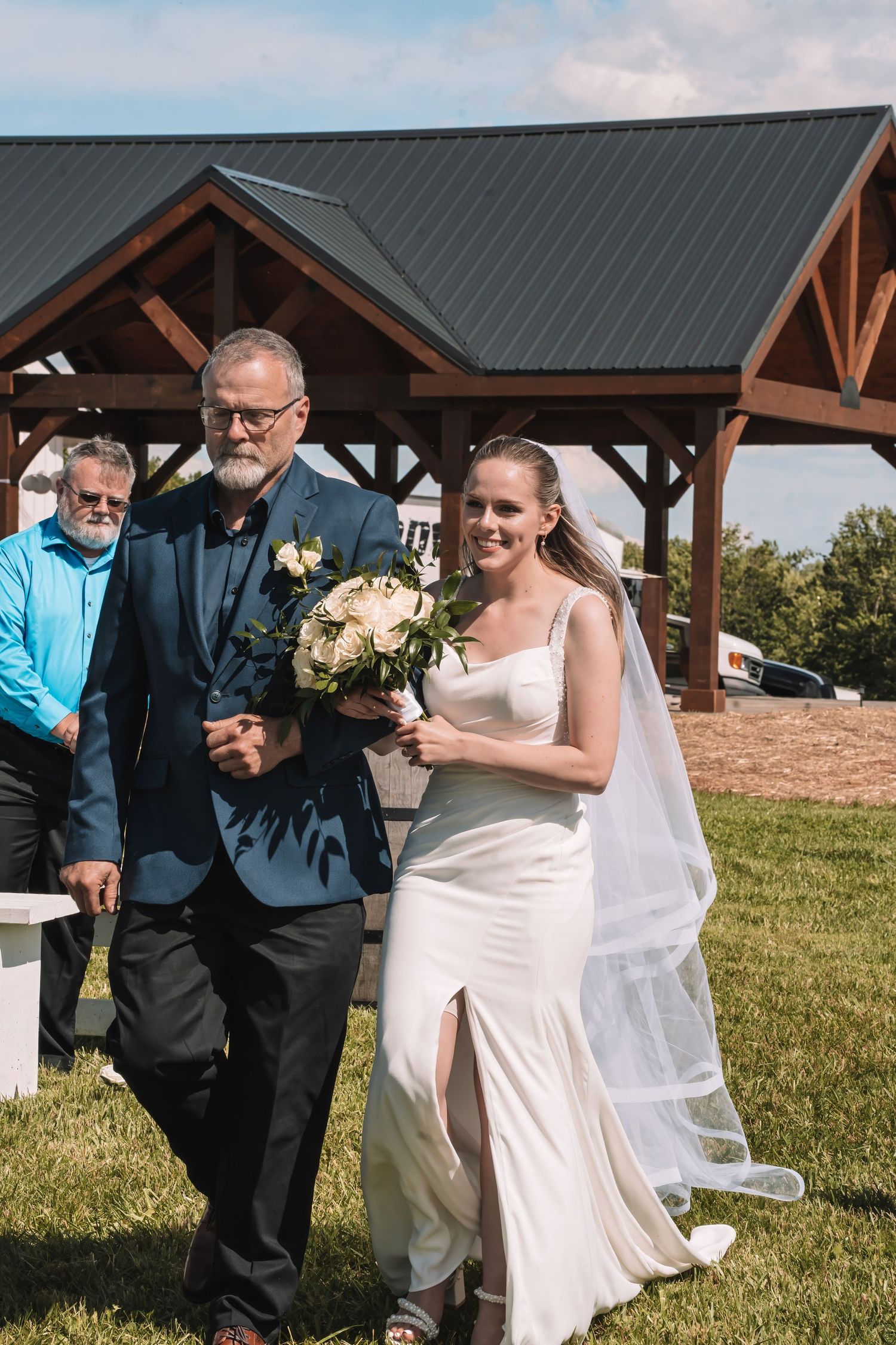 A bride in a white wedding dress with a flowing veil walks down a grass aisle under an outdoor pavilion.