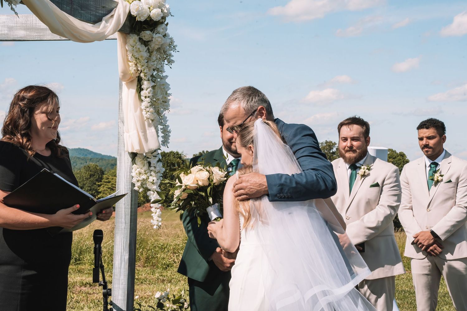 An emotional outdoor wedding ceremony taking place under a white draped arbor with mountains in the background.