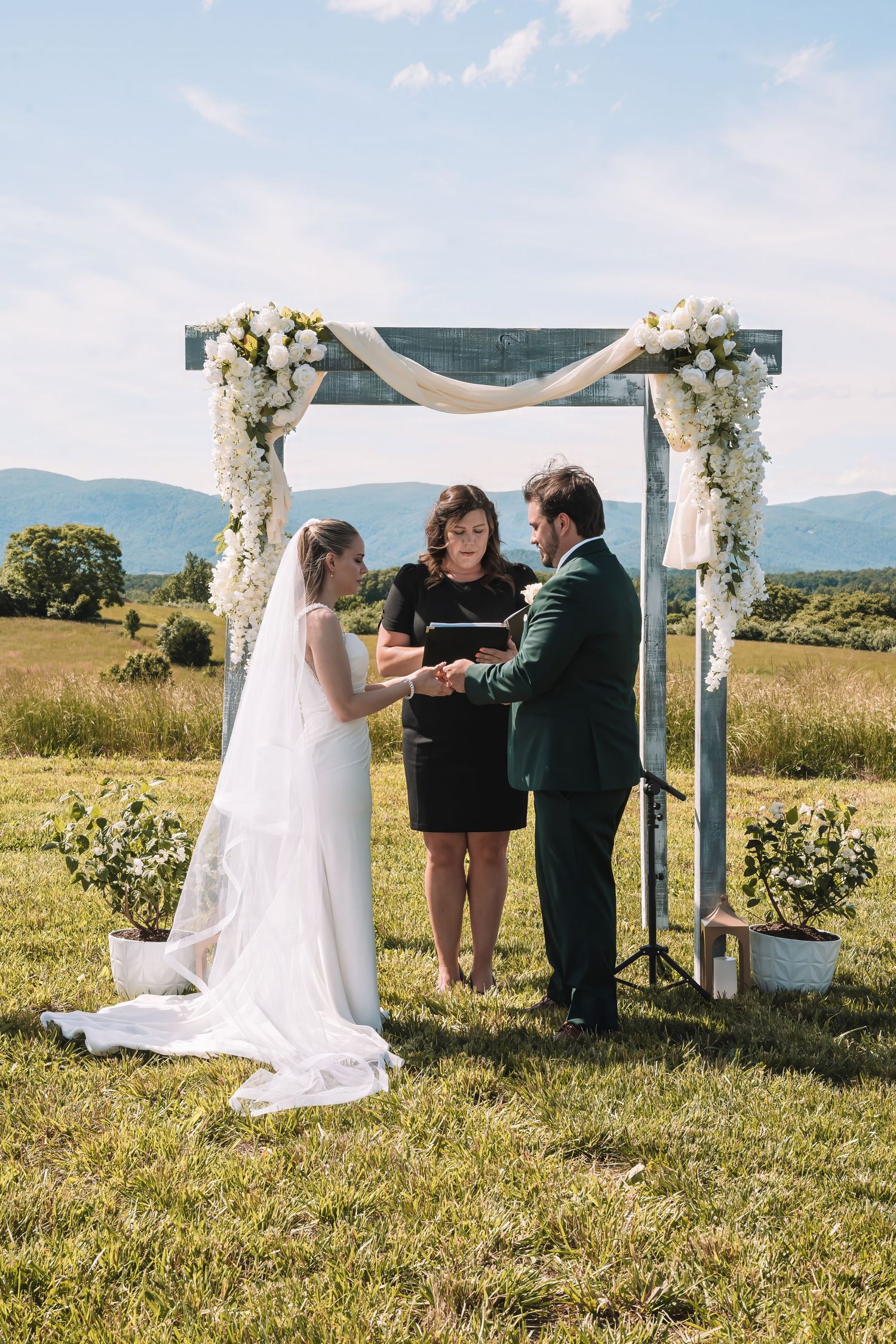 An outdoor wedding ceremony beneath a white floral arch with mountains in the background on a sunny summer day.