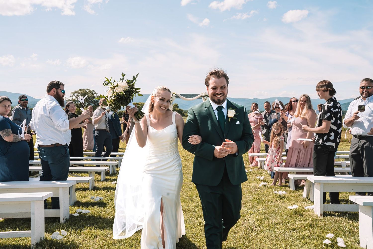 Wedding ceremony on a hilltop meadow with white benches and guests gathered on a sunny day.