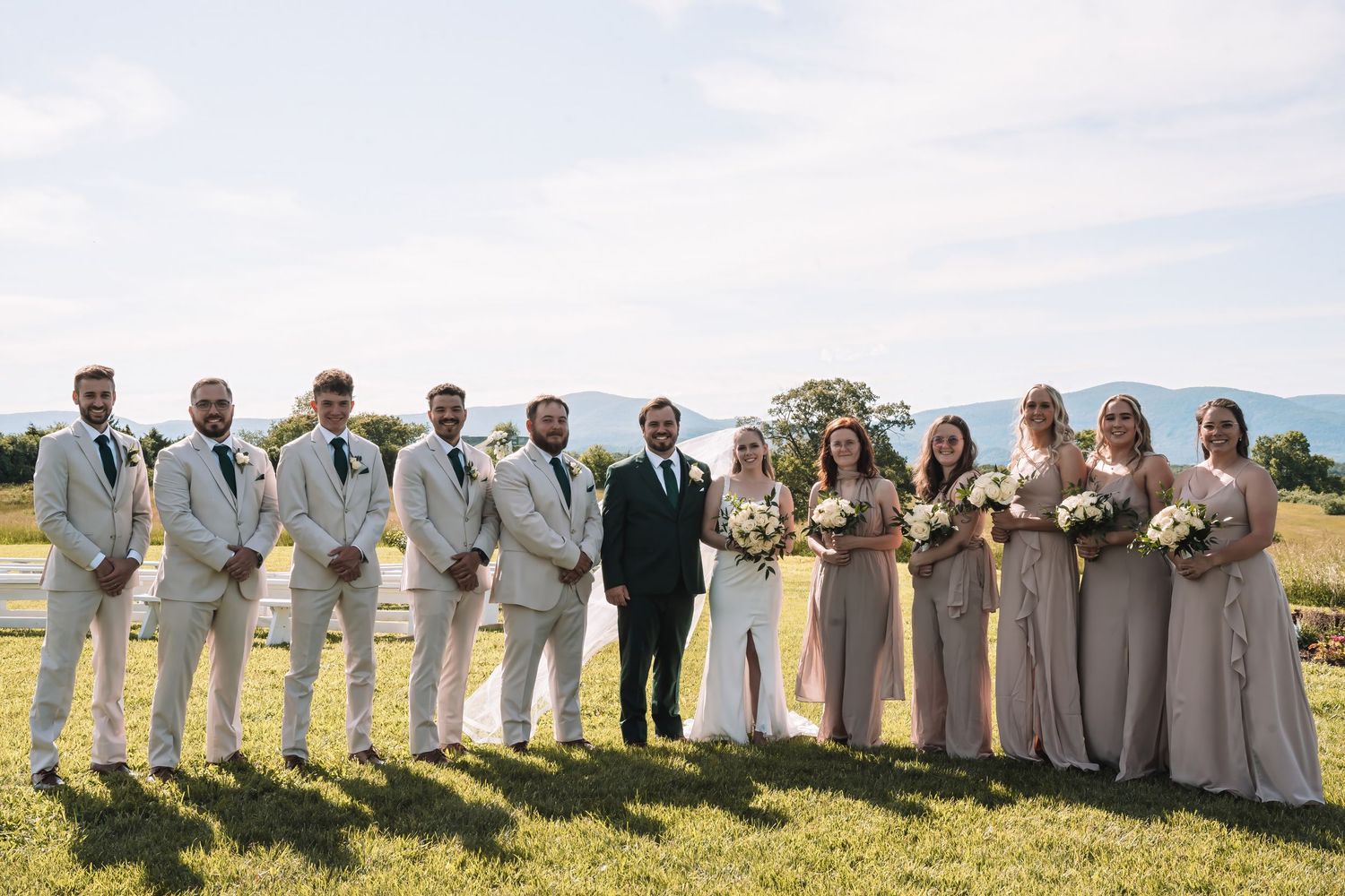 Wedding party in light gray suits and neutral dresses poses together on a grassy hilltop with ocean view backdrop.