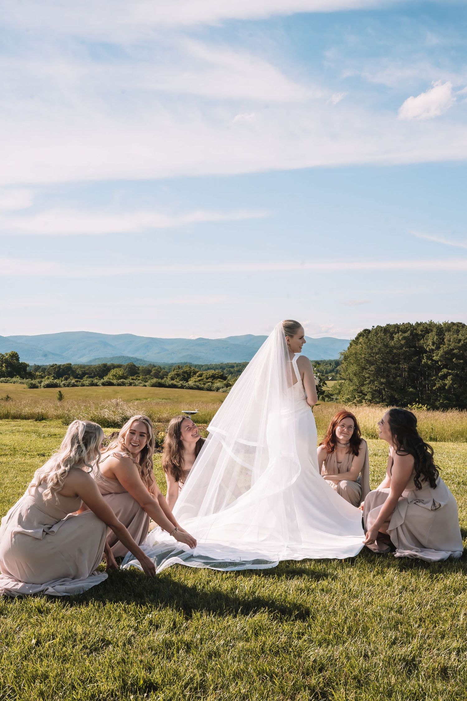 Bridesmaids help arrange a flowing white wedding dress train in a sunny meadow with mountains in the background.