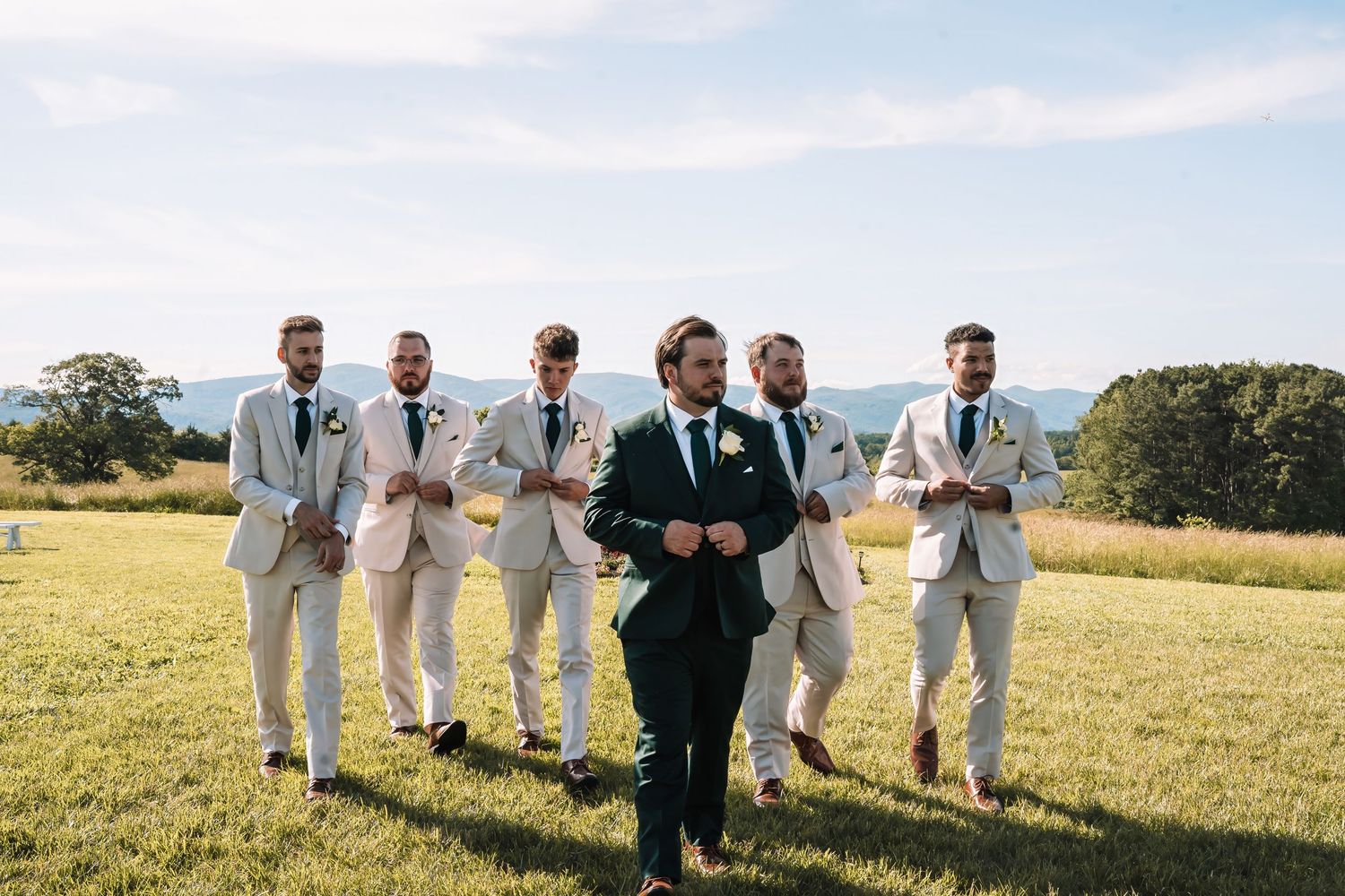 Groomsmen in light gray suits walk across sunny field with person in dark suit during outdoor wedding ceremony.