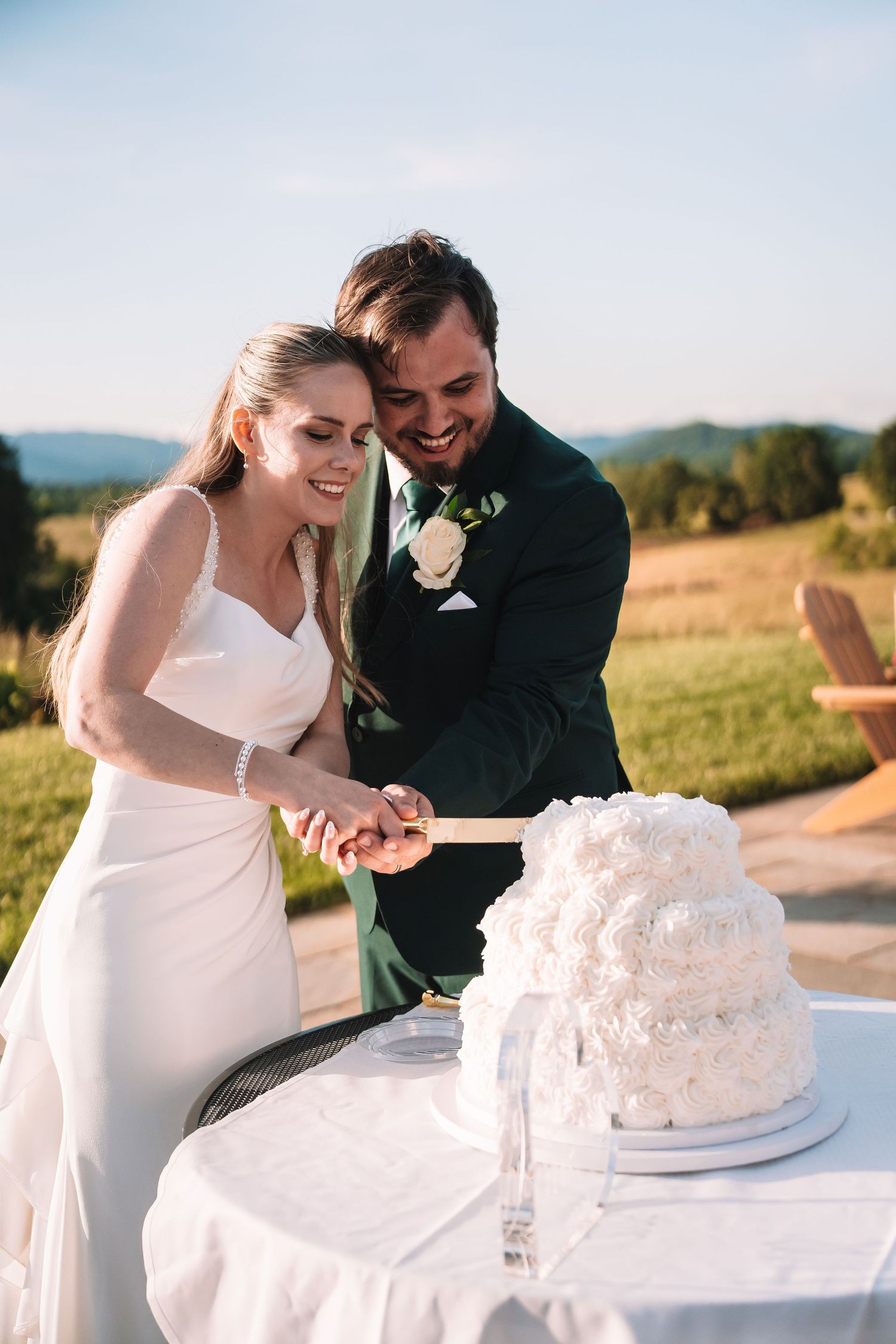 A newlywed couple cuts into their elegant white wedding cake outdoors at a scenic countryside venue during sunset.