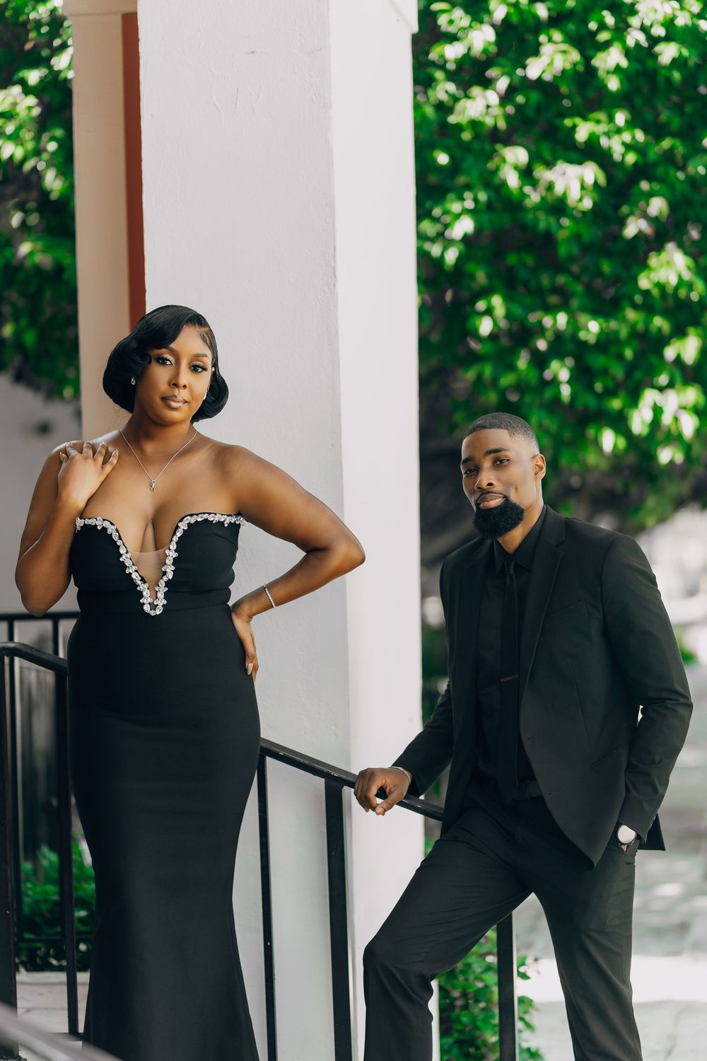 Elegant couple in all black formal attire pose on outdoor steps with lush greenery in the background.