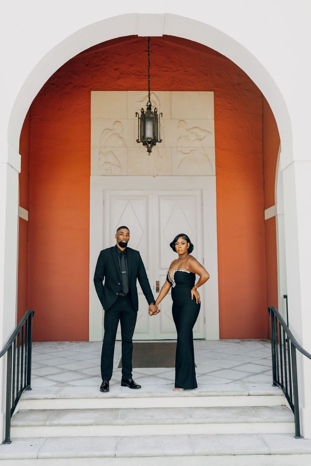 Elegant couple in formal black attire poses beneath a terracotta-colored archway with white trim and hanging lantern.