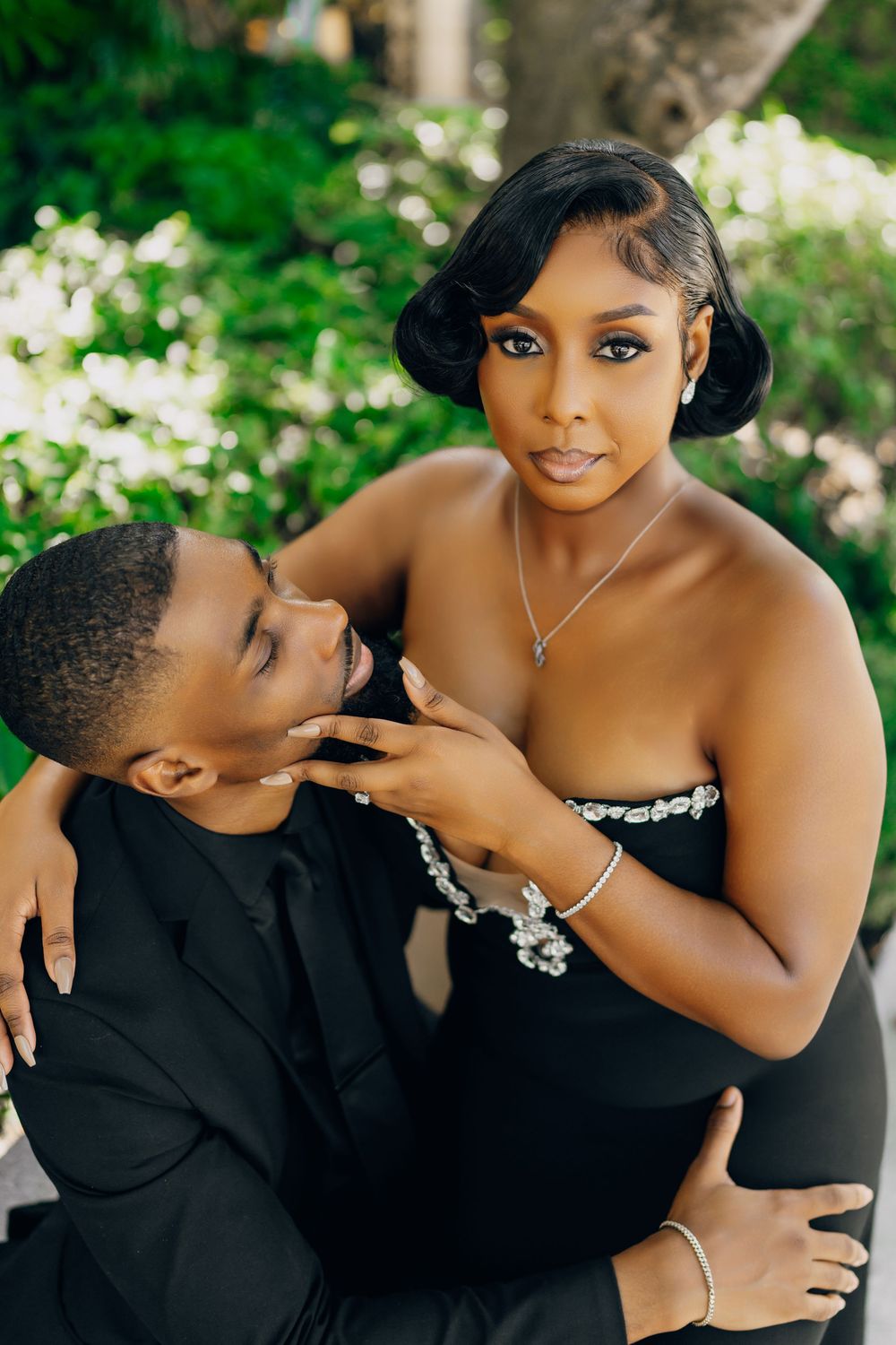 Elegant couple in formal black attire pose together outdoors with lush greenery in a romantic engagement photo.
