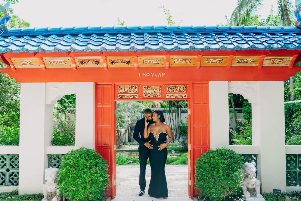 Couple stands in embrace at ornate red and white Chinese garden entrance with palm trees in background.